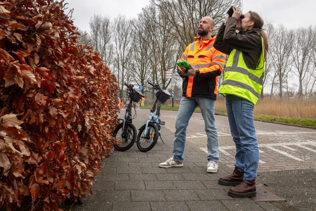 Twee mensen op straat in hesjes kijken met verrekijker naar huizen