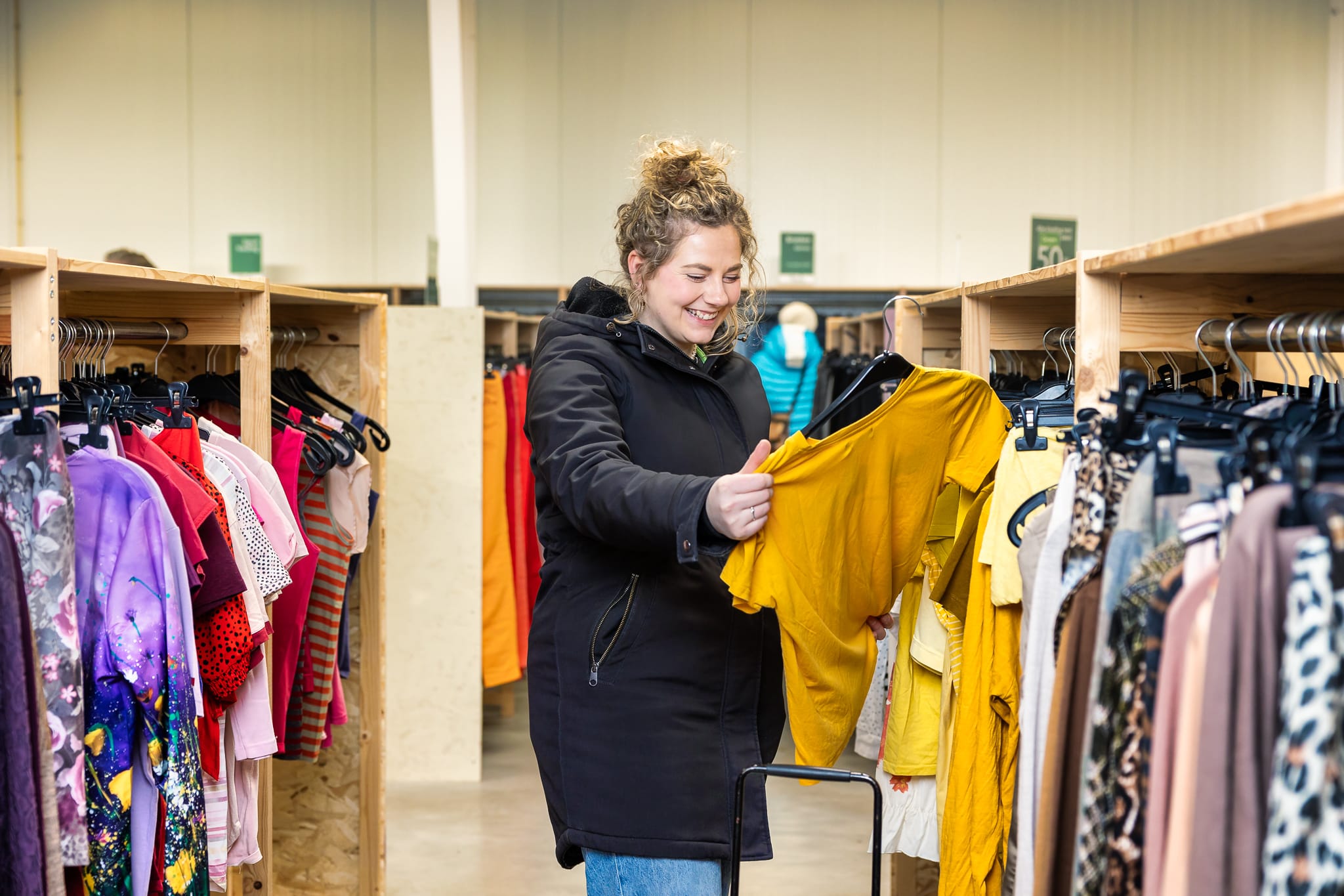 Een vrouw bekijkt een gele top die zij uit een rek heeft gehaald in een kringloopwinkel. Foto: RataPlan Alkmaar