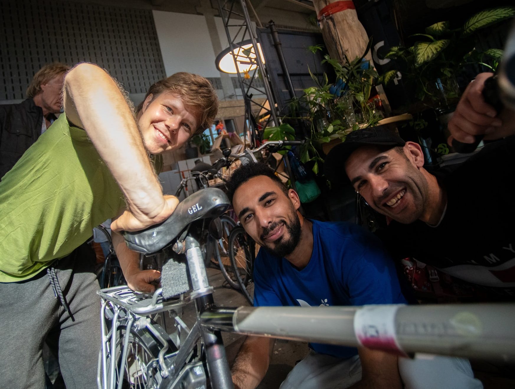 3 mannen kijken en lachen in de camera, 1 houdt zijn hand op het zadel van een fiets