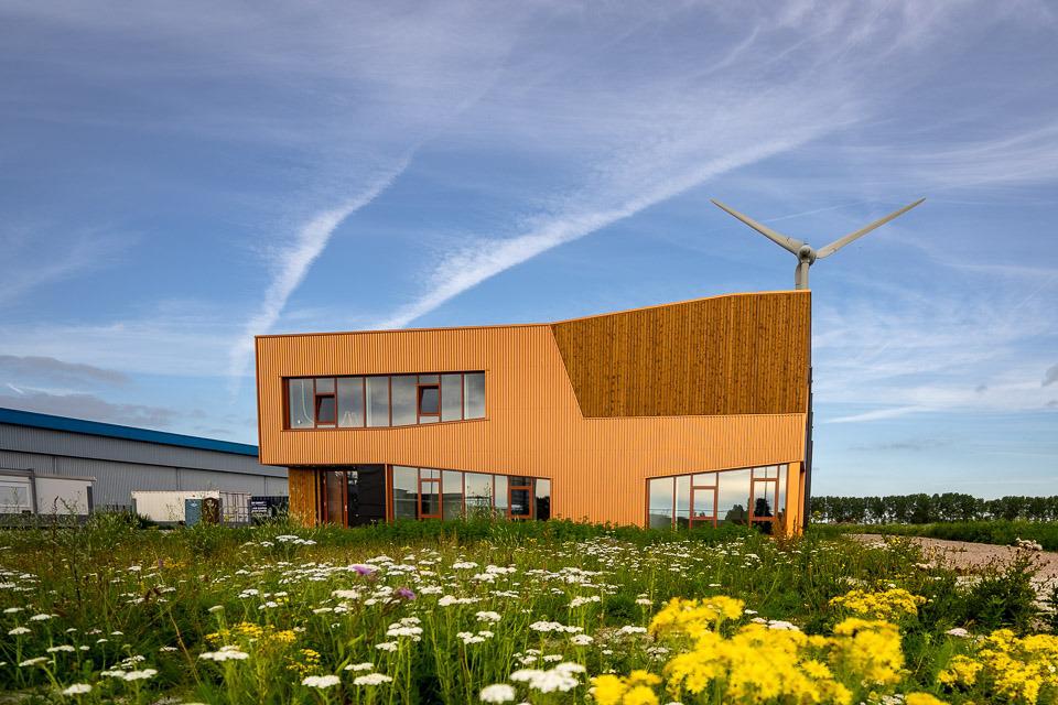 Terracotta kleurig gebouw van investa tegen een blauwe lucht met op de achtergrond een windmolen en op de voorgrond een veld met gras en gele en witte bloemen. 