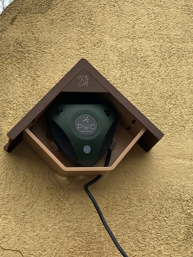 Een meetkastje van Trackvogel dat lijkt op een vogelhuisje. Het hangt aan een gele muur. 