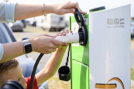 Een hand sluit een stekker aan op een laadpaal. De persoon houdt met de andere hand de stekkerdop open. Naast de paal een kind en op de achtergrond auto's en een weg. 