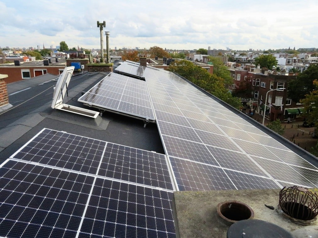 Zonnepanelen op een groot en lang dak. Foto genomen vanaf het dak. Uitzicht op omliggende wijk. 