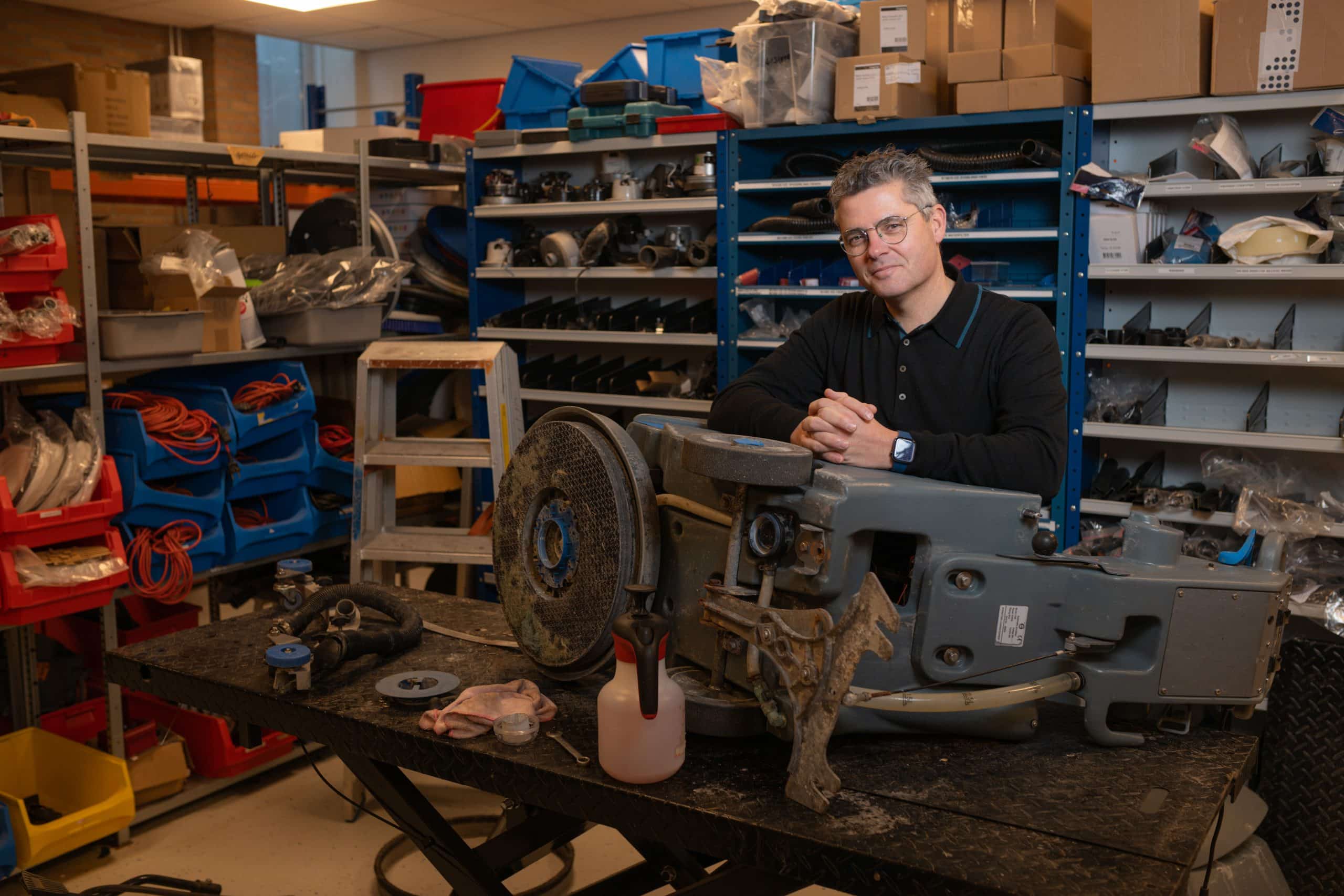 Man in werkplaats leunt op machine. Op de achtergrond veel gereedschappen en onderdelen. 